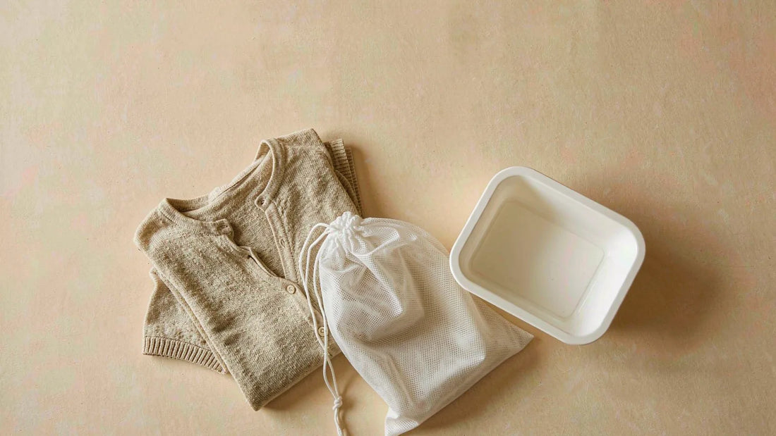 A folded oatmeal knit top beside a white mesh laundry bag and small basin on a warm cream background