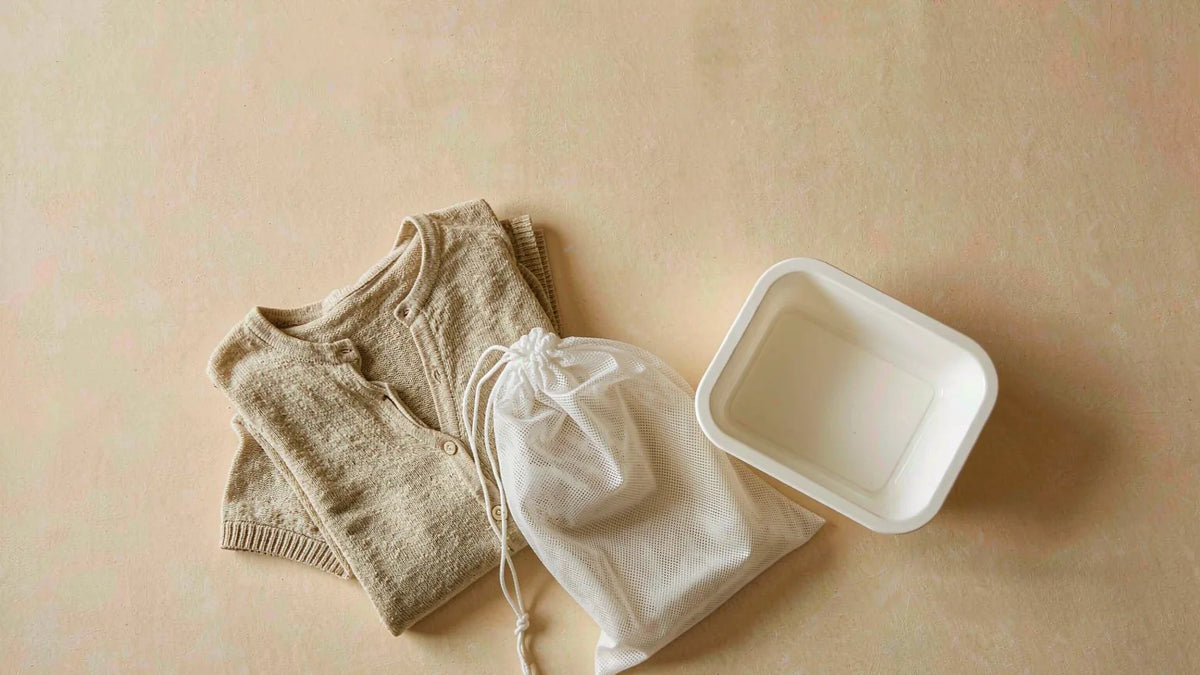 A folded oatmeal knit top beside a white mesh laundry bag and small basin on a warm cream background