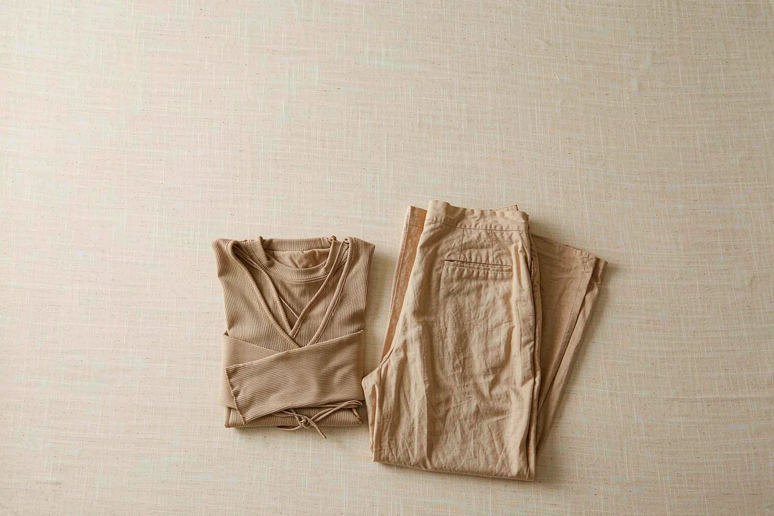 A single capsule outfit laid flat: a folded knit top beside folded wide-leg trousers on a warm cream background