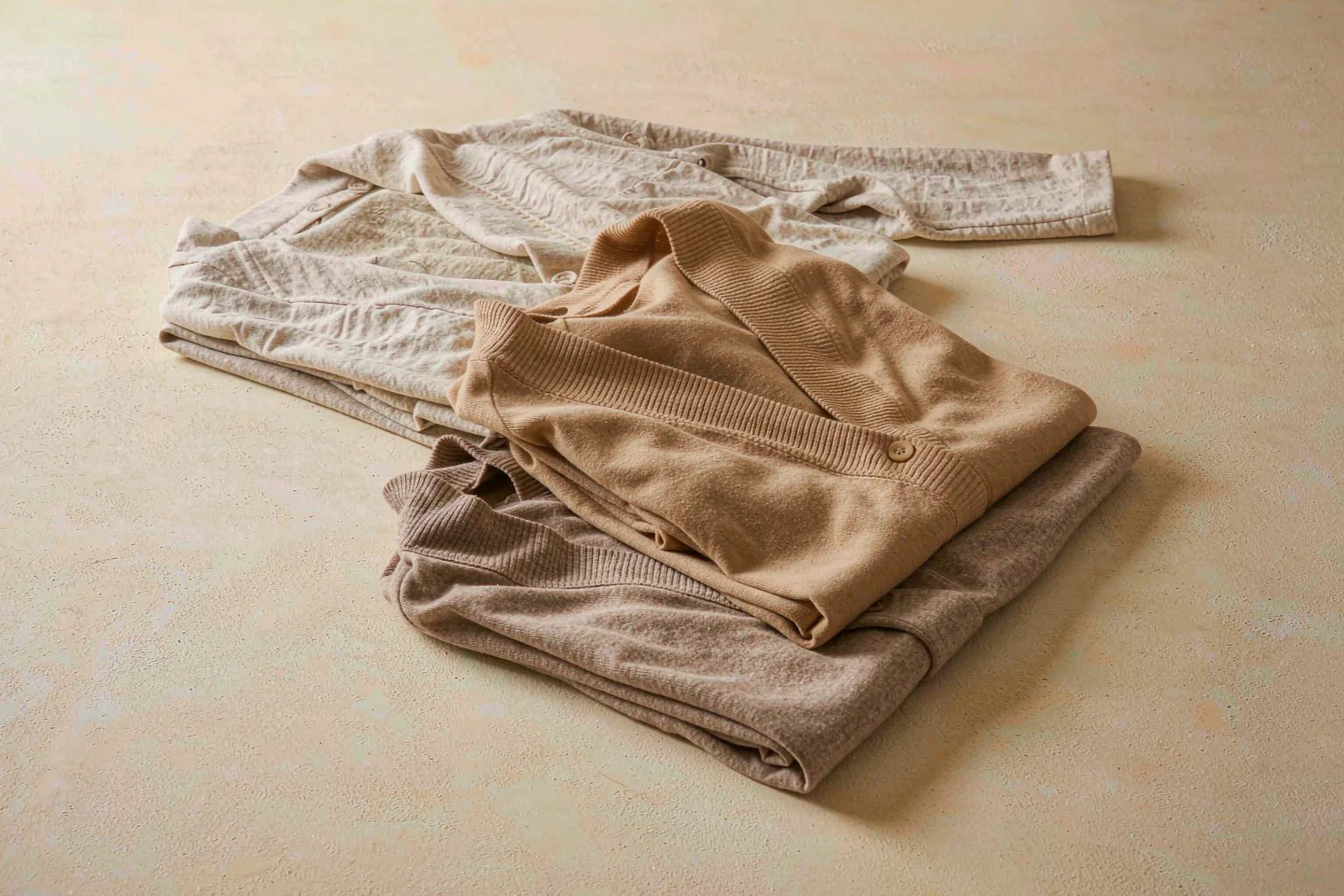 A neat stack of folded knit tops in oatmeal, camel and warm taupe earth tones on a cream surface