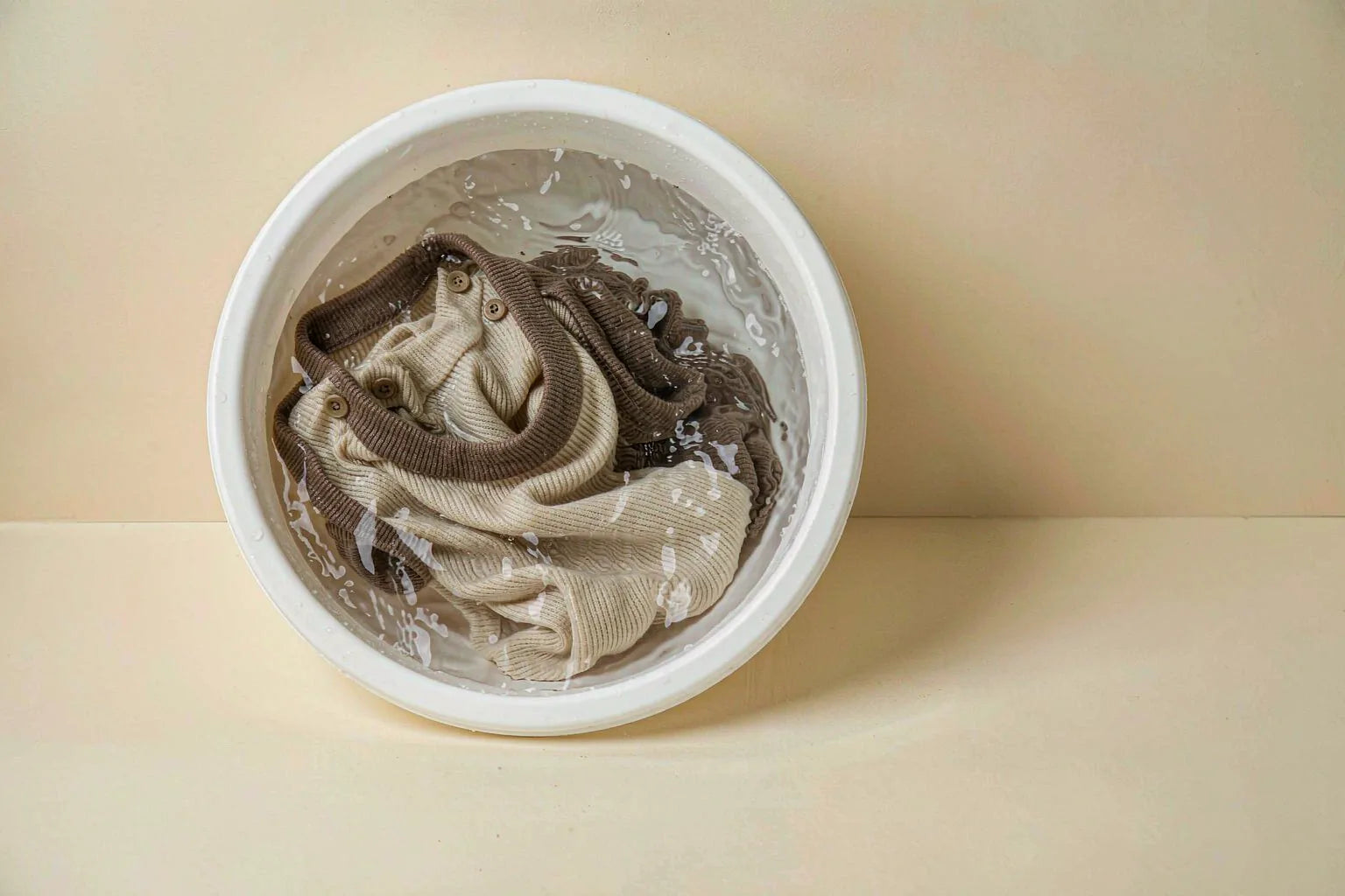 A folded knit top soaking in a white basin of clear water on a warm cream surface