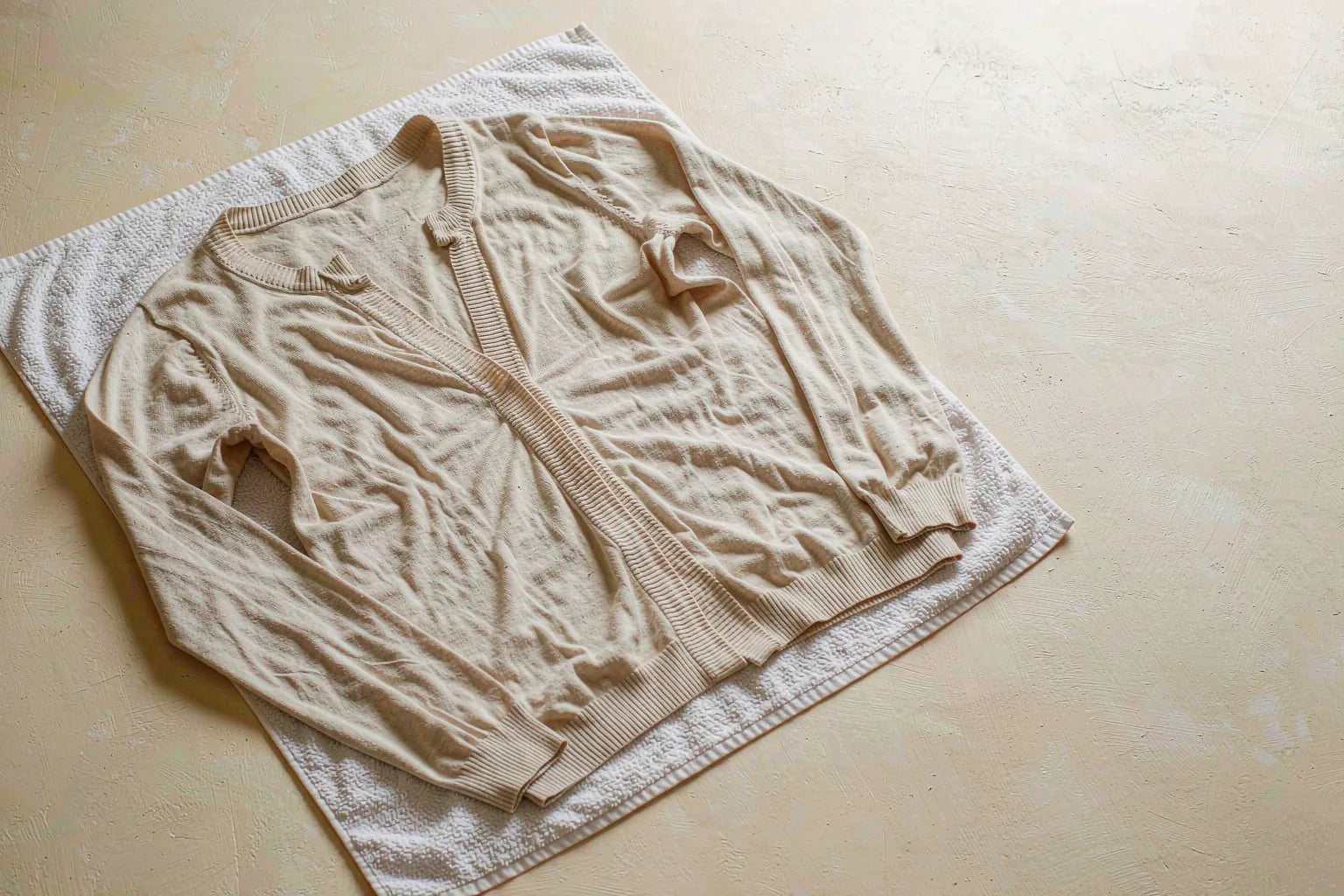 An oatmeal knit top laid flat on a white towel to dry, sleeves straightened, on a warm cream surface