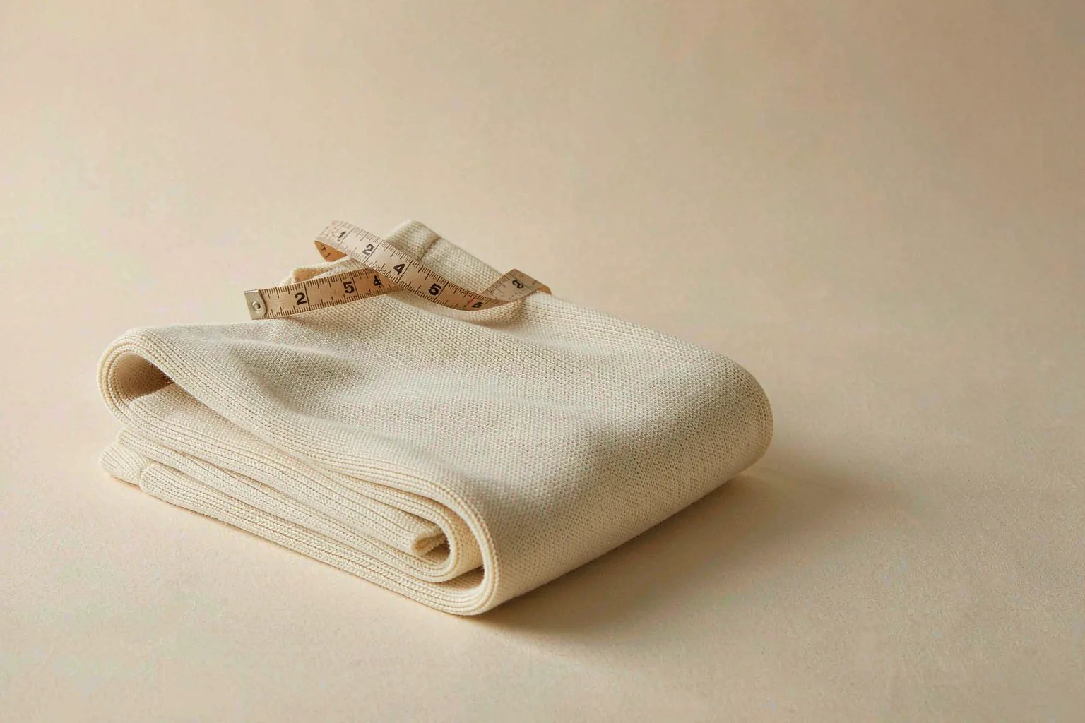 A soft beige measuring tape draped over a folded cream knit top on a warm off-white surface
