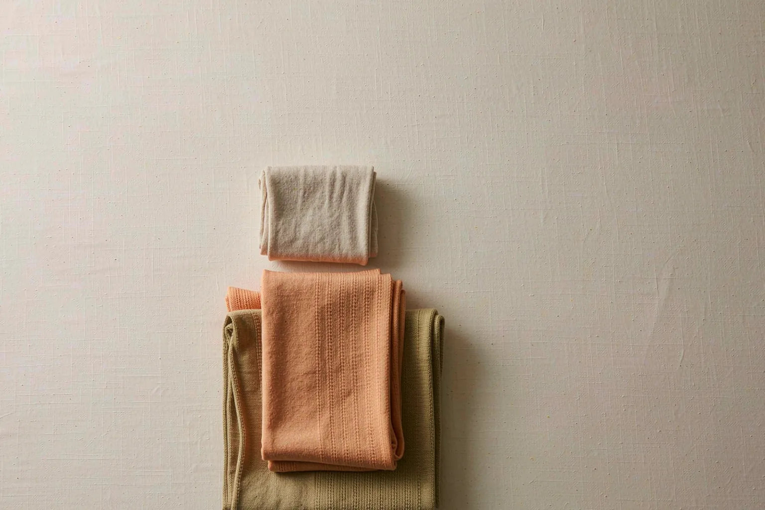 Flat lay of a capsule quiet-luxury wardrobe with folded neutral knits in apricot, oatmeal and khaki arranged on cream linen