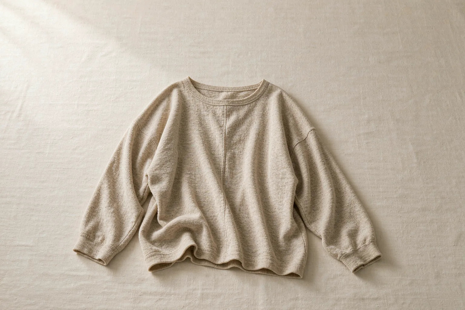 An oatmeal relaxed-fit knit top laid on a flat surface with softly draped sleeves showing natural drape on a warm cream background