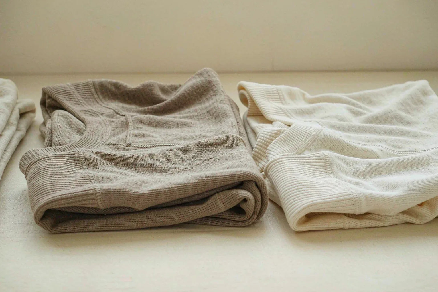 Two folded knit tops in oatmeal and warm ivory placed side by side on a cream background, one slightly larger than the other